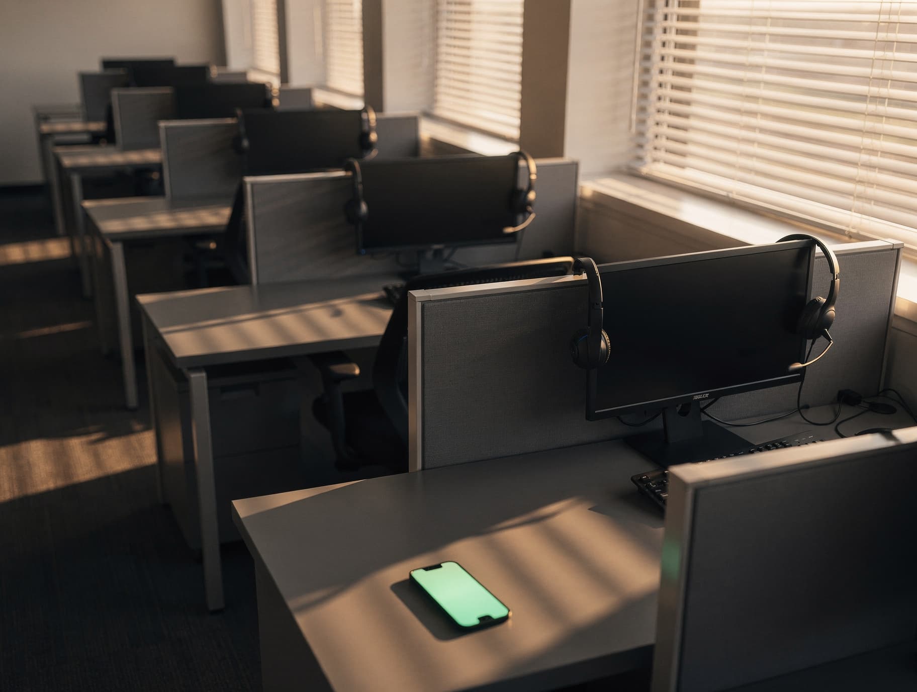 Fileira de posições de atendimento vazias em um call center, com headsets pendurados nos monitores desligados e luz de fim de tarde entrando pela janela